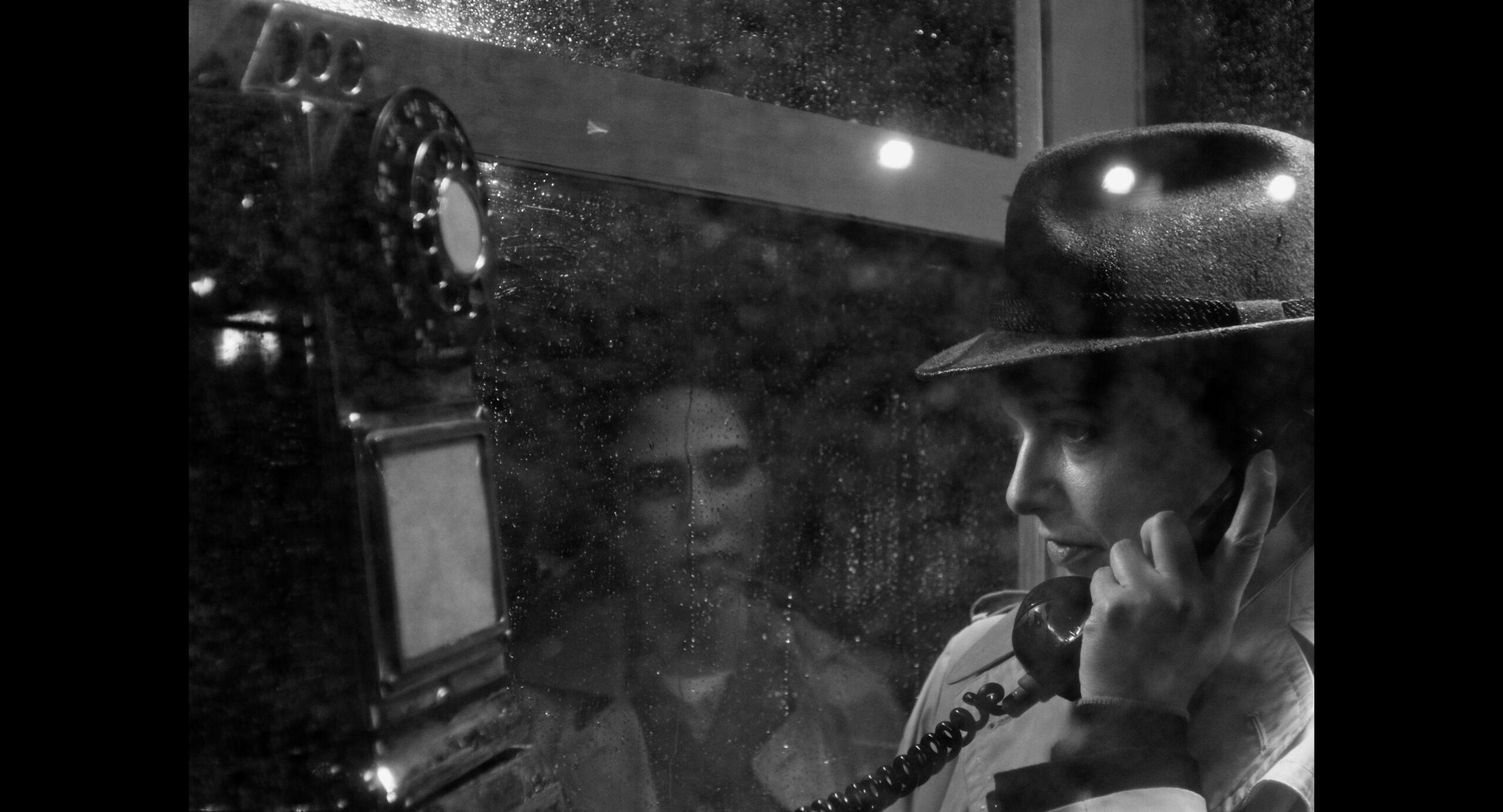 Butch talks nervously in a phone booth while a zombie-like woman stares at them while standing in the rain.