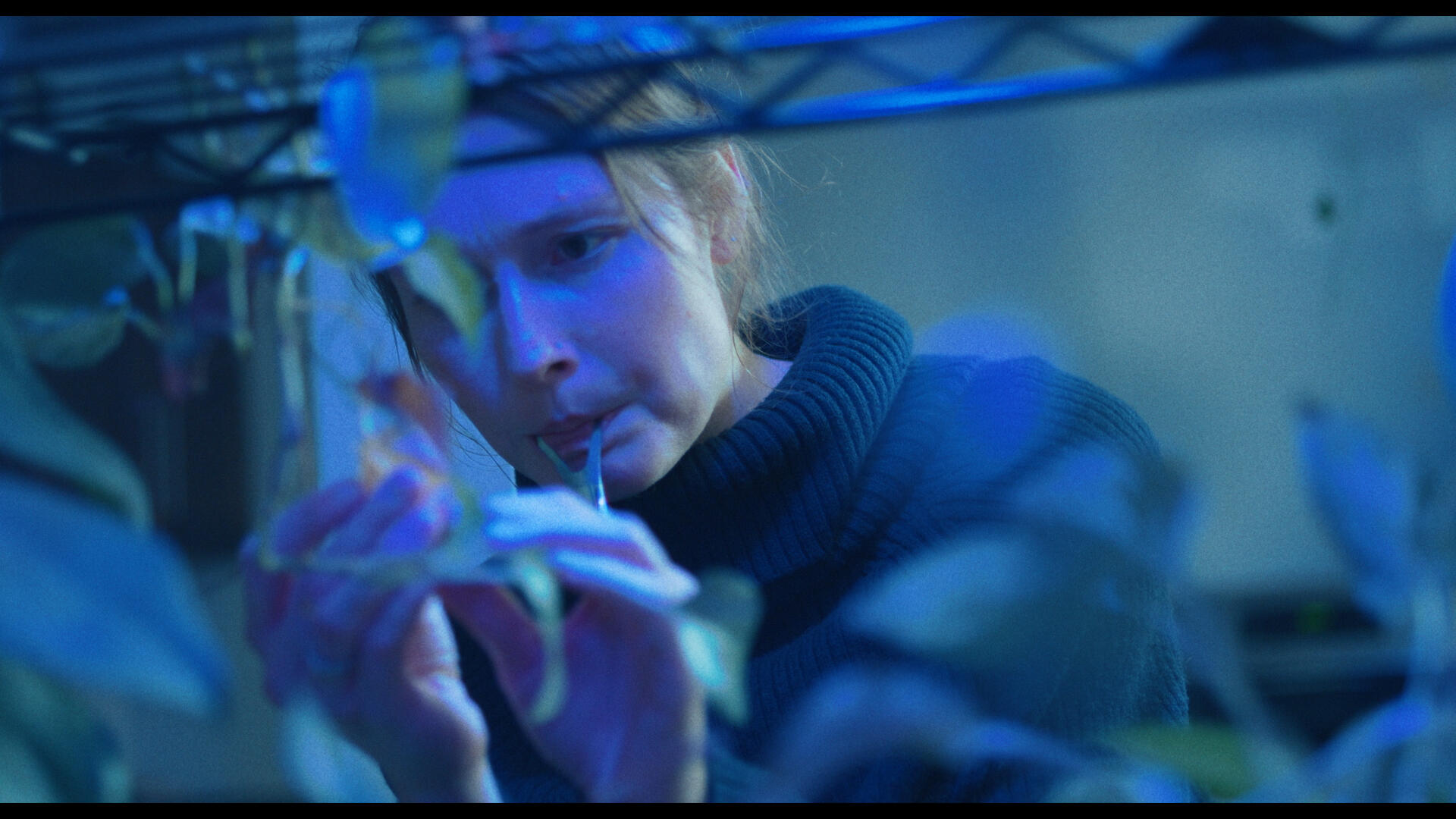 The protagonist of Working For The Weekend tends to some plants on a wire rack, illuminated by a vivid blue light. She's holding a tongue scraper in her mouth.